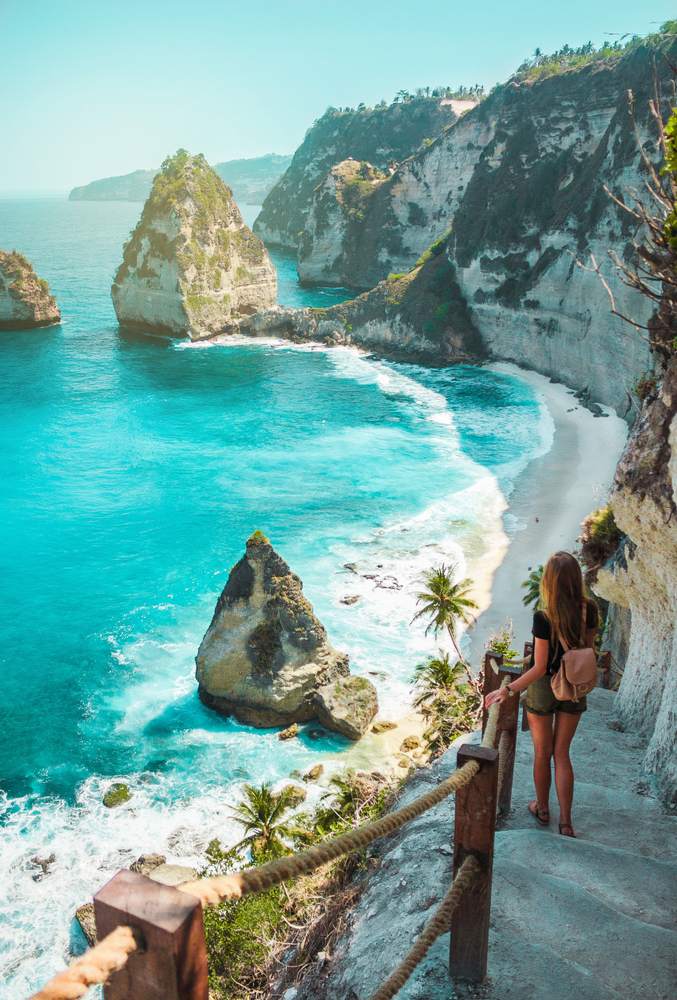 Una ragazza da viaggio con zaino e pantaloncini sull'oceano, scogliere e sfondo spiaggia tropicale. Nusa Penida, Indonesia. Silhouette femminile