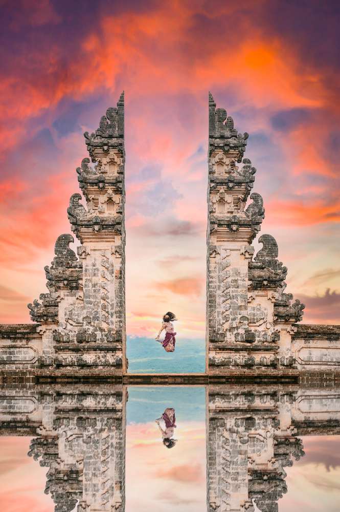 Una ragazza salta davanti alla Porta del Cielo con il suo riflesso nell'acqua durante uno splendido tramonto. Bali, Indonesia.