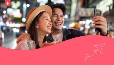 Coppia asiatica di turisti zaino in spalla che si scatta un selfie e registra un video blog mentre mangia cibo da una bancarella nel mercato notturno affollato di Yaowarat Road, Bangkok.