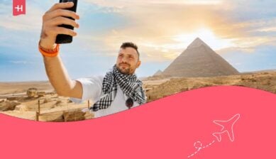 Un ragazzo, davanti alla piramide, alza il cellulare e si scatta un selfie con le dita davanti all'obiettivo.