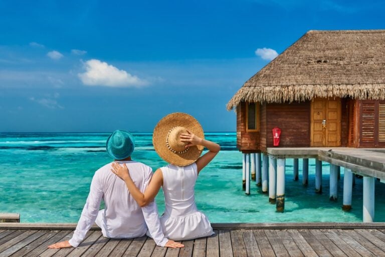 Coppia in abito bianco su un molo in una spiaggia tropicale vicino a una villa sull'acqua alle Maldive.