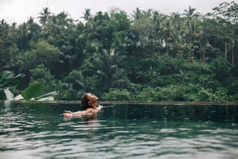 Una donna felice che si gode la calda pioggia tropicale che le cade addosso mentre nuota in una piscina a sfioro con vista sulla giungla a Ubud, Bali