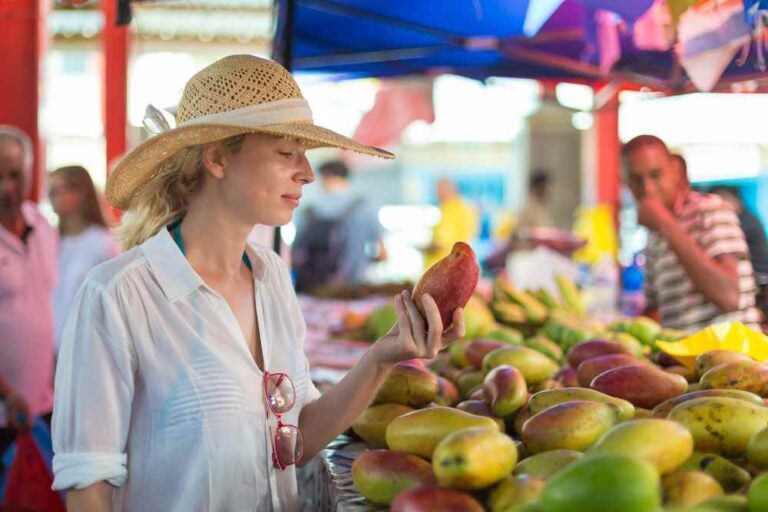 Una viaggiatrice che indossa un’elegante tunica bianca in stile coloniale e un cappello mentre acquista frutta tropicale fresca al tradizionale mercato alimentare di Victoria, nelle Isole Seychelles.