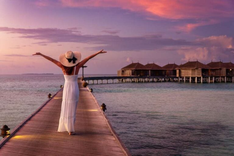 Una donna sorridente, con un abito bianco e un cappellino, cammina lungo un molo affacciato sull’oceano turchese delle Isole Maldive durante un tramonto dai colori vivaci