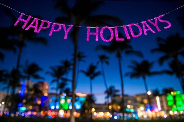Un colorato messaggio di “Buone Feste” appeso su uno striscione scintillante sopra una vista notturna sfocata dello skyline di Miami Beach, con luci al neon e palme.