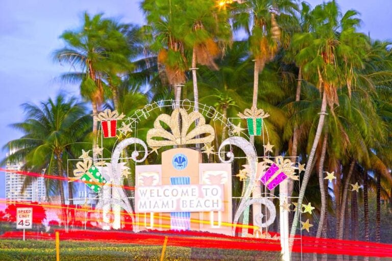 Cartello di benvenuto a Miami Beach con decorazioni natalizie e palme al tramonto, con il traffico in movimento.