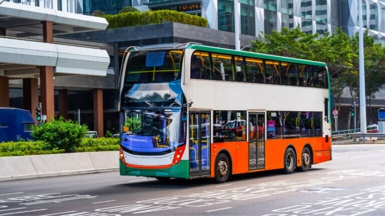 Een wit-oranje Citybus dubbeldekker die door een straat in Hong Kong rijdt met moderne wolkenkrabbers op de achtergrond.