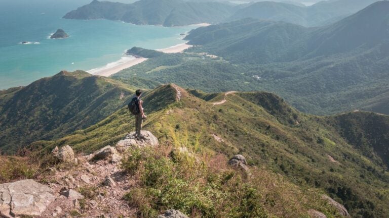 Een wandelaar met een rugzak op een rotsachtige bergtop met uitzicht op het turquoise water en de witte zandstranden van Sai Kung, Hong Kong.