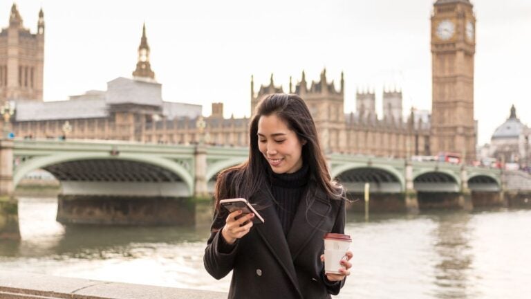 Een jonge vrouw die lachend op haar smartphone kijkt met een koffie in haar hand voor de Big Ben en Westminster Bridge in Londen, een van de beste stedentrips Europa.