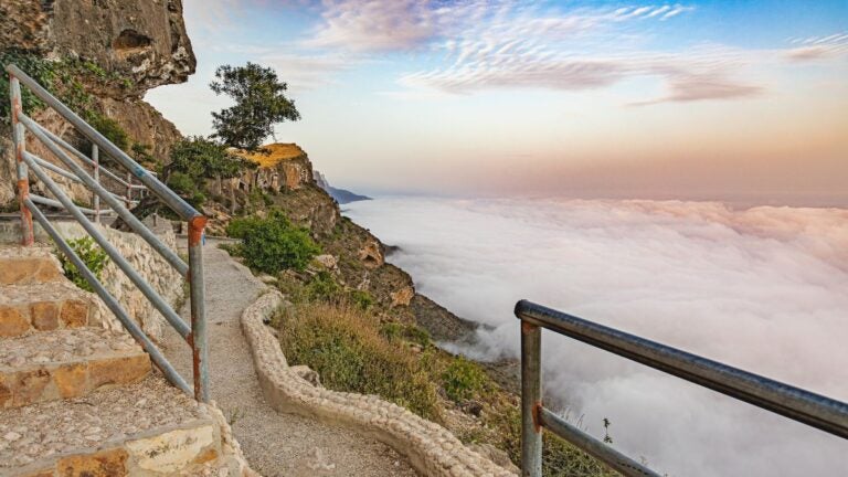 Een wandelpad langs de rand van Jebel Shams in Oman, uitkijkend over een indrukwekkend wolkendek tussen de bergen bij zonsondergang