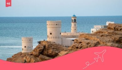 Historische witte wachttorens en de Al Ayjah-vuurtoren met een kenmerkende blauwe koepel in Sur, Oman, uitkijkend over de turquoise zee met ruige rotsen op de voorgrond.