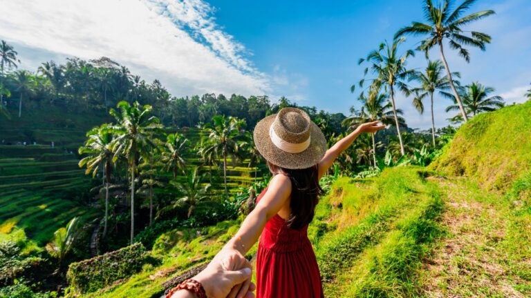 Een persoon houdt de hand vast van een vrouw in een rode jurk en hoed, kijkend over de groene rijstvelden op Bali, Indonesië.