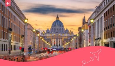 Zonsondergangzicht op de Sint-Pietersbasiliek in Rome, gezien vanaf een verlichte straat met stadsverkeer op de voorgrond.