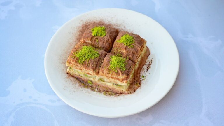 Stukjes Griekse baklava met laagjes deeg en fijngemalen pistachenoten op een wit bord.