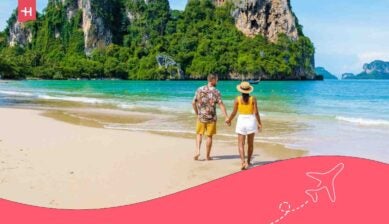 Koppel wandelt hand in hand langs een tropisch strand in Thailand, met turquoise water en kalkstenen kliffen op de achtergrond.