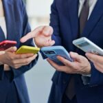 ビジネススーツを着た男女がそれぞれのスマートフォンを手に取りながら意見交換する様子。カラフルなスマホケースが目を引くビジネスシーン。