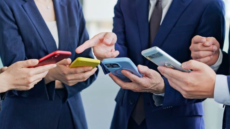 ビジネススーツを着た男女がそれぞれのスマートフォンを手に取りながら意見交換する様子。カラフルなスマホケースが目を引くビジネスシーン。