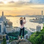 香港のビクトリア・ピークから、夕暮れの高層ビル群とビクトリア湾を背景にスマートフォンで撮影している様子