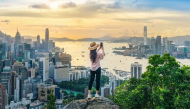 香港のビクトリア・ピークから、夕暮れの高層ビル群とビクトリア湾を背景にスマートフォンで撮影している様子