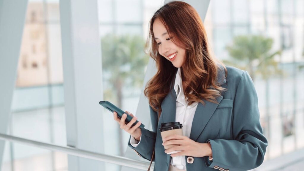 スマートフォンを見ながら笑顔でコーヒーを持つ女性ビジネスパーソン