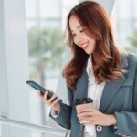 スマートフォンを見ながら笑顔でコーヒーを持つ女性ビジネスパーソン