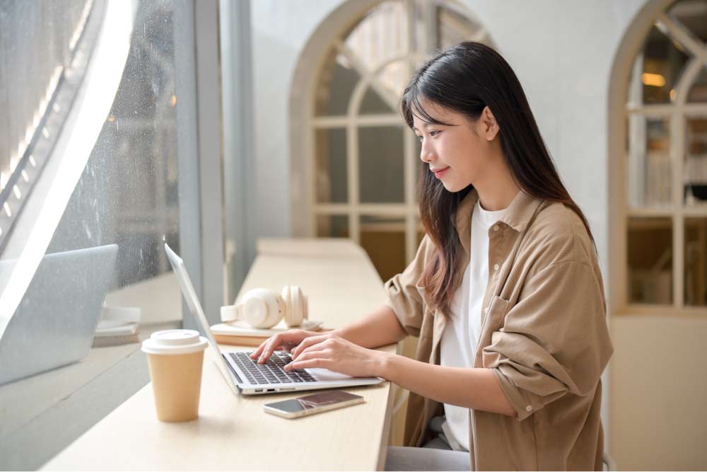 カフェの窓際でノートパソコンを使って作業する女性。リラックスした雰囲気の中で集中してタイピングしている様子。