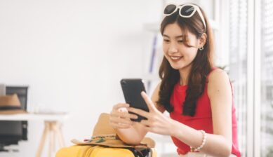 黄色いスーツケースに座り、スマホで旅行準備をする女性