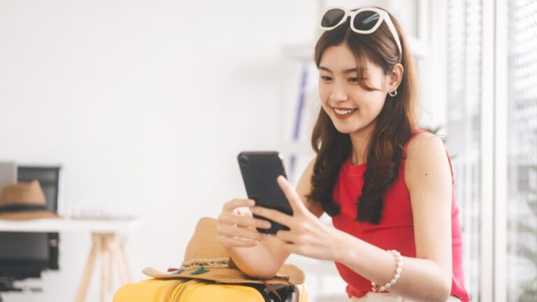 黄色いスーツケースの隣に座り、スマホで旅行準備をする女性