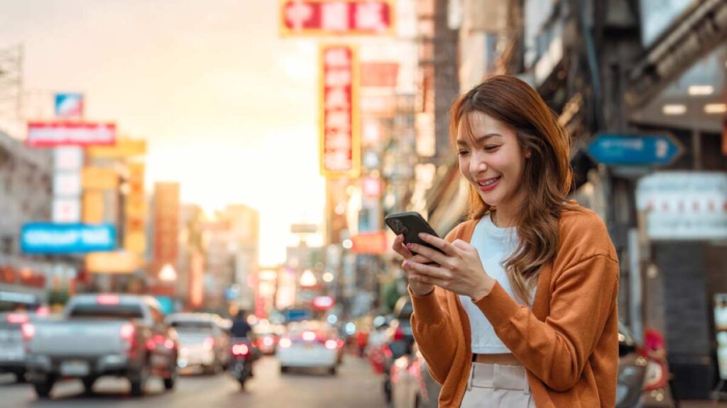 中国の繁華街を背景に、スマートフォンを操作しながら微笑む女性。都市の喧騒とテクノロジー利用の様子が伝わるシーン。