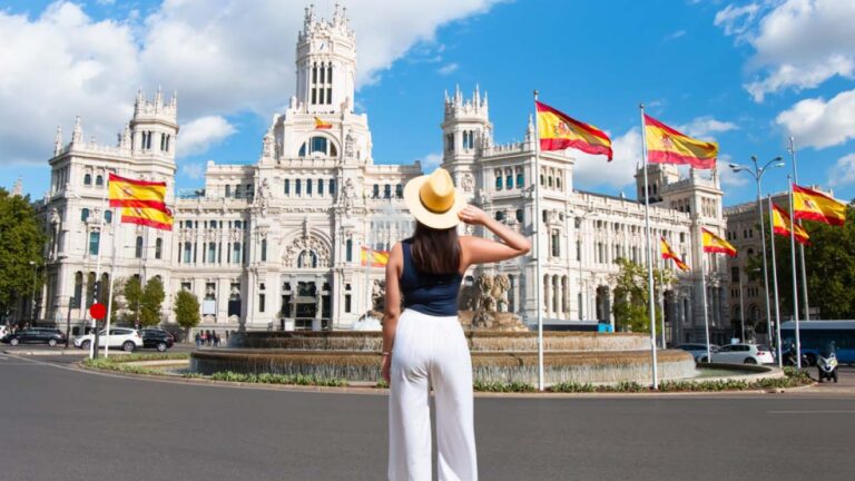 スペイン・マドリードのシベーレス宮殿を背景に、スペイン国旗がはためく広場に立つ女性旅行者。白いパンツと帽子を身に着け、観光を楽しむ姿が印象的。