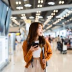 空港の出発ロビーでスマートフォンを手にフライト情報を確認する女性旅行者。