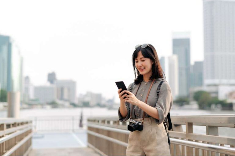 川沿いの遊歩道でスマートフォンを見る旅行中の女性。首からカメラを下げ、都会の高層ビルを背景に笑顔を見せている。