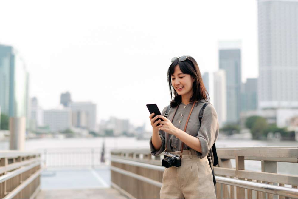 川沿いの遊歩道でスマートフォンを見る旅行中の女性。首からカメラを下げ、都会の高層ビルを背景に笑顔を見せている。