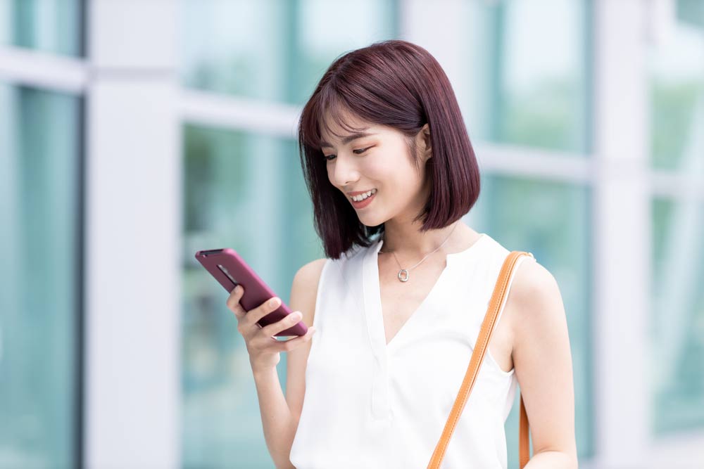 スマートフォンを見ながら笑顔を浮かべるアジア系の若い女性。明るい屋外で、ガラス張りの建物を背景にリラックスした表情を見せる日常の一コマ。
