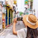 麦わら帽子をかぶった女性が、シンガポールのハジ・レーンでカラフルなストリートアートをスマホで撮影している様子。