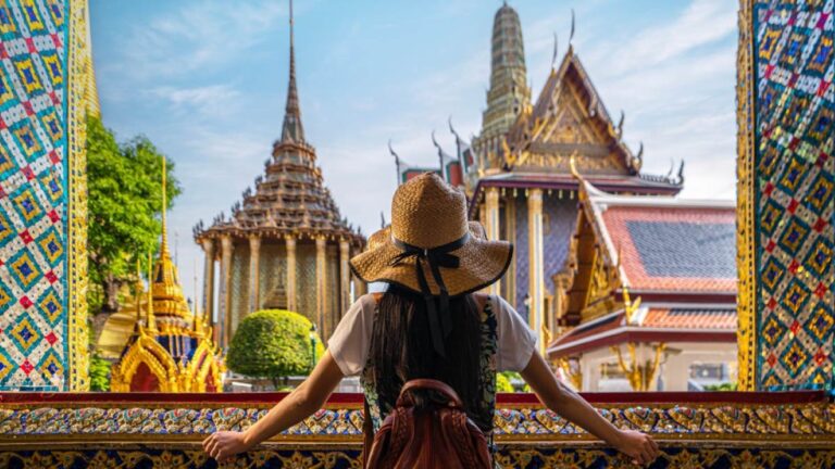 タイ・バンコクの王宮「グランドパレス」を眺める女性旅行者。装飾が美しい寺院と鮮やかな瓦屋根が広がる歴史的建造物を背景に、帽子とリュックを身に着けた後ろ姿。