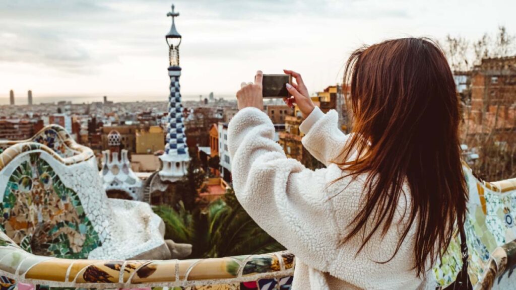 バルセロナのグエル公園で街並みを撮影する観光客の女性。ガウディ建築のモザイクベンチと塔、遠景に広がる都市の風景が特徴。