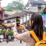 中国の伝統的な街並みを背景に、スマートフォンで写真を撮る黄色いリュックを背負った若い女性の後ろ姿。赤い提灯や木造建築が並ぶ観光地の雰囲気が漂っている。