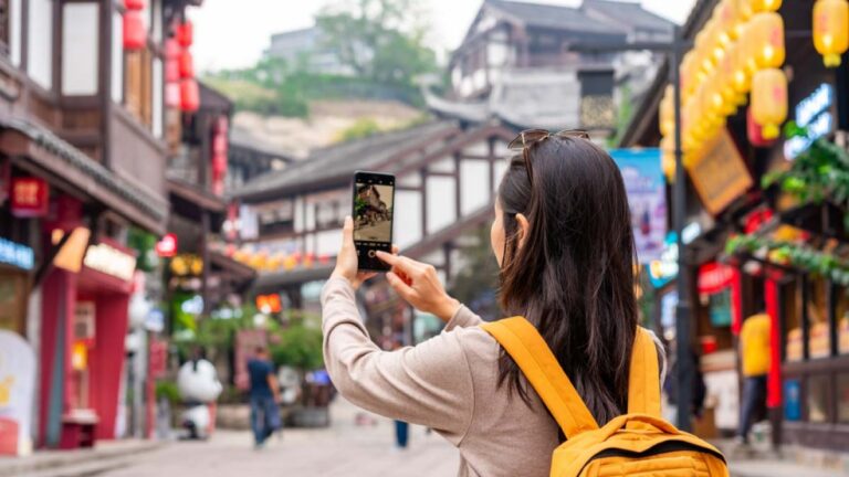 中国の伝統的な街並みを背景に、スマートフォンで写真を撮る黄色いリュックを背負った若い女性の後ろ姿。赤い提灯や木造建築が並ぶ観光地の雰囲気が漂っている。