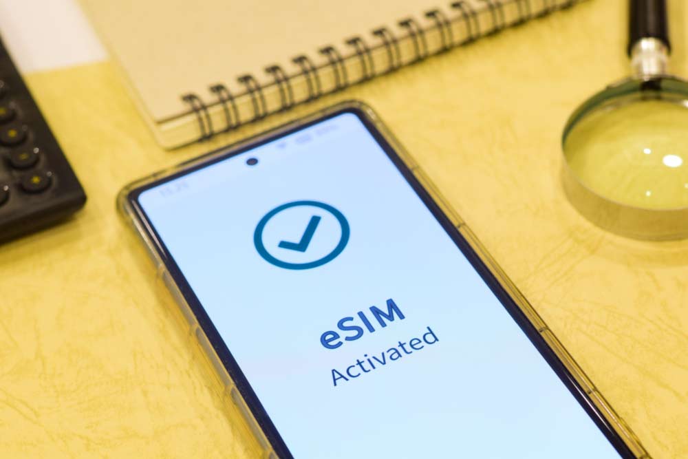 スマートフォンの画面に「eSIM Activated」と表示されている様子。eSIMの設定が完了した状態を示しており、周囲にはノートや虫眼鏡などが置かれている。