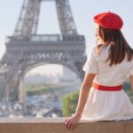 エッフェル塔を背景に赤いベレー帽と白いワンピースを着た女性が座って景色を眺めている後ろ姿。