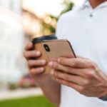 屋外でスマートフォンを操作しながらコーヒーを持つ男性の手元。カジュアルな白いシャツとスマートウォッチが印象的。