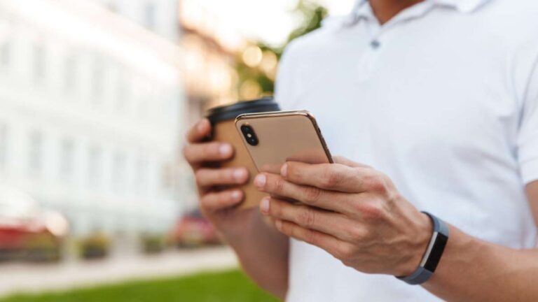 屋外でスマートフォンを操作しながらコーヒーを持つ男性の手元。カジュアルな白いシャツとスマートウォッチが印象的。