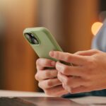 グリーンのスマホケースを付けたスマートフォンを操作する人物の手元、デスク上にはノートパソコンと書類が置かれている。