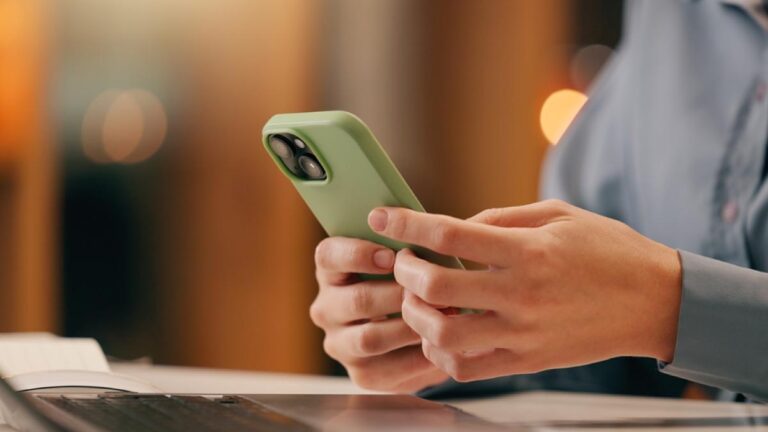 グリーンのスマホケースを付けたスマートフォンを操作する人物の手元、デスク上にはノートパソコンと書類が置かれている。