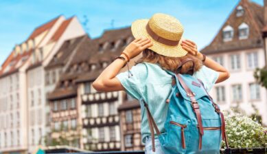 ヨーロッパの旧市街を背景に、麦わら帽子とバックパック姿で街並みを眺める旅行者の後ろ姿