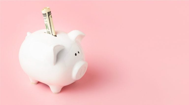 ピンクの背景に置かれた白い貯金箱に、紙幣が差し込まれている。