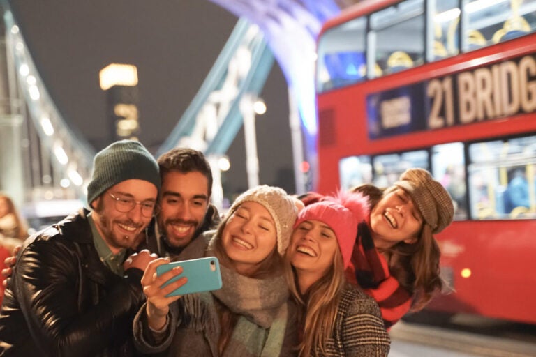 イギリス・ロンドンのタワーブリッジ近くで、赤い二階建てバスを背景に笑顔で自撮りをする若者グループ。