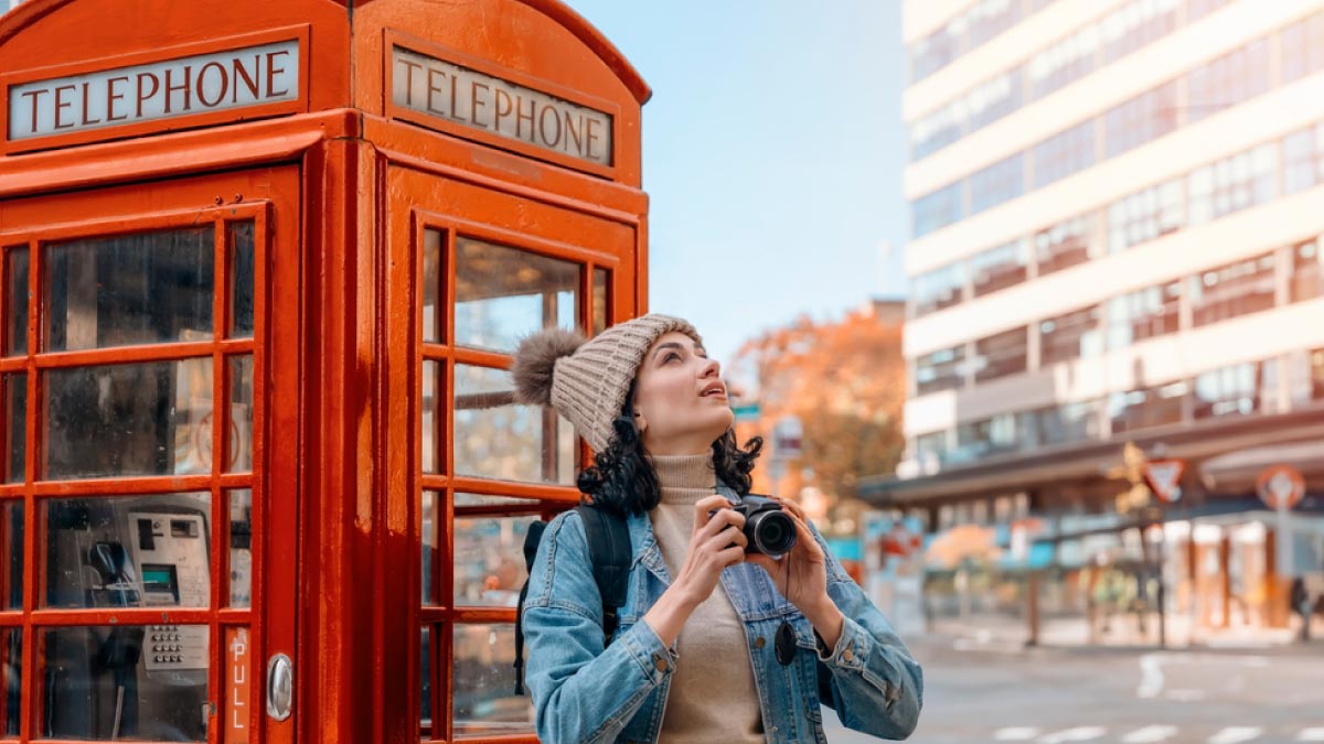 イギリス・ロンドンの赤い電話ボックスの前で、カメラを持って空を見上げる女性旅行者。秋の街並みに映える観光シーン。