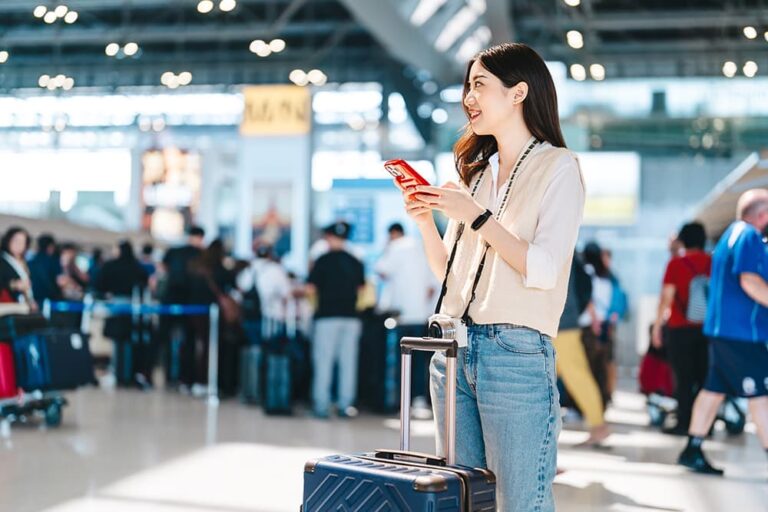 混雑する空港ターミナルで、スーツケースの横に立ちスマートフォンで通信設定を確認している女性旅行者の様子